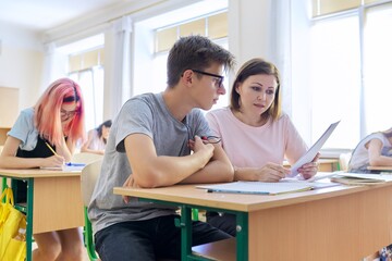 Fototapeta premium Middle-aged woman teacher sitting at desk with teenage student teaches