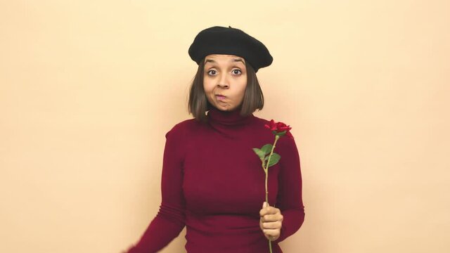 Young latin woman with boina holding rose shrugs shoulders and open eyes confused