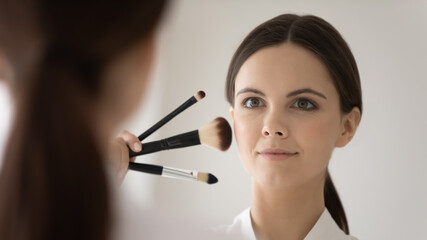 Preparing to make beauty. Happy inspired young woman facialist reflected in mirror choosing foundation brush from makeup kit collection starting doing professional visage creating visualizing image