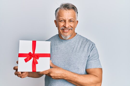 Middle age grey-haired man holding gift looking positive and happy standing and smiling with a confident smile showing teeth
