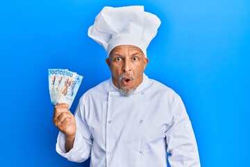 Middle age grey-haired man wearing professional cook uniform holding swiss franc banknotes scared and amazed with open mouth for surprise, disbelief face