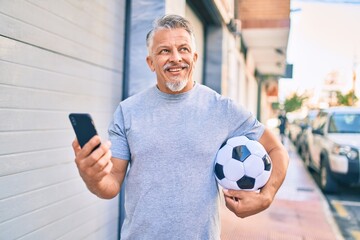 Middle age hispanic grey-haired man using smartphone and holding soccer ball at the city.