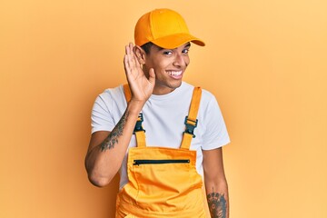 Young handsome african american man wearing handyman uniform over yellow background smiling with hand over ear listening an hearing to rumor or gossip. deafness concept.