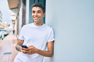 Young latin man smiling happy using smartphone leaning on the wall at the city.