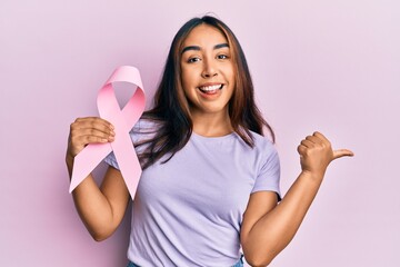 Young latin woman holding pink cancer ribbon pointing thumb up to the side smiling happy with open mouth