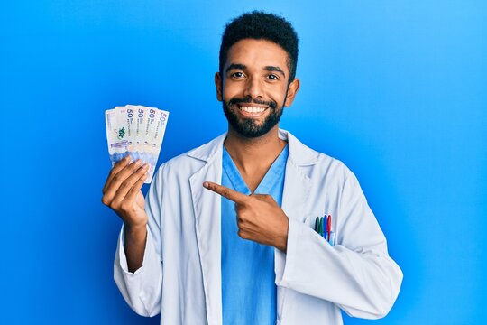 Handsome young hispanic doctor man holding colombian pesos banknotes scared and amazed with open mouth for surprise, disbelief face