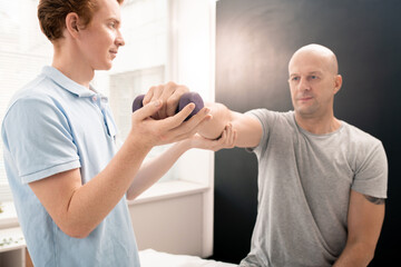 Obraz premium Young physiotherapist helping patient lifting and stretching arm with dumbbell