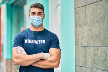 Young hispanic volunteer man with arms crossed wearing medical mask at the city.