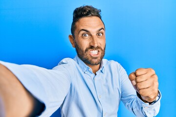 Handsome man with beard taking a selfie photo with smartphone annoyed and frustrated shouting with anger, yelling crazy with anger and hand raised