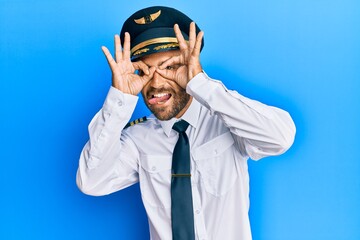 Handsome man with beard wearing airplane pilot uniform doing ok gesture like binoculars sticking tongue out, eyes looking through fingers. crazy expression.