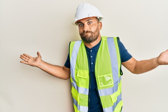 Handsome man with beard wearing safety helmet and reflective jacket clueless and confused with open arms, no idea and doubtful face.