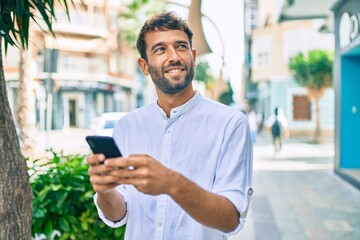 Handsome man with beard wearing casual white shirt on a sunny day smiling happy outdoors using smartphone