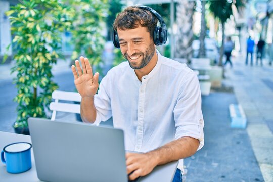 Handsome man with beard wearing casual white shirt on a sunny day working using laptop and wearing headphones and waving to videocall at cafeteria