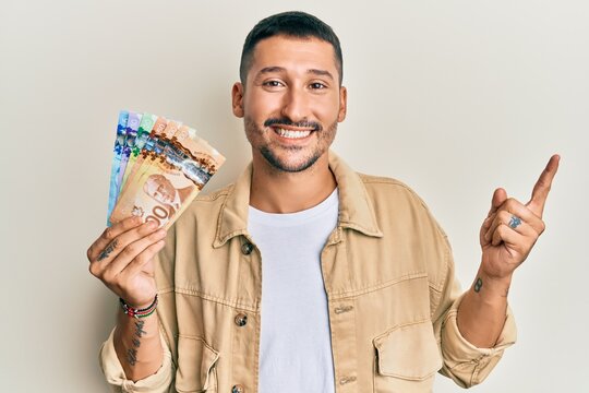 Handsome man with tattoos holding canadian dollars smiling happy pointing with hand and finger to the side