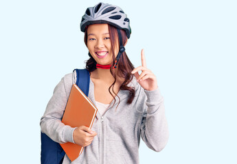 Young beautiful chinese girl wearing student backpack and bike helmet holding book surprised with an idea or question pointing finger with happy face, number one