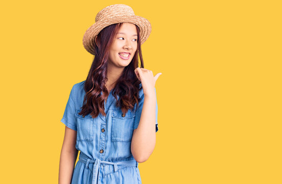 Young beautiful chinese girl wearing summer hat smiling with happy face looking and pointing to the side with thumb up.