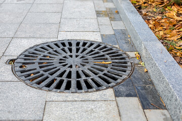 manhole cover round black metal lattice on a city park pedestrian sidewalk paved with granite gray tiles with a stone border close-up view autumn season with fallen yellow leaves, nobody.