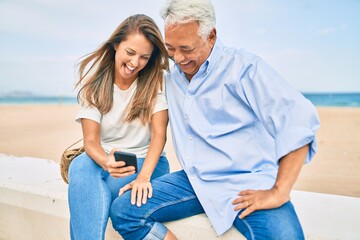 Middle age hispanic couple using smartphone sitting on the bench at the beach.