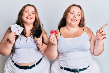 Plus size caucasian sisters woman holding poker chips and cards smiling happy pointing with hand and finger to the side