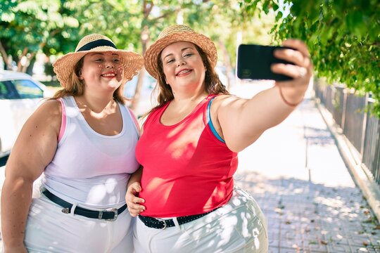 Two plus size overweight sisters twins women smiling taking a selfie picture with the phone outdoors on a sunny day