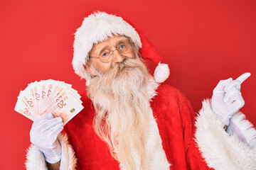 Old senior man with grey hair and long beard wearing santa claus costume holding israel shekels smiling happy pointing with hand and finger to the side