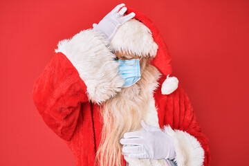 Old senior man wearing santa claus costume wearing safety mask surprised with hand on head for mistake, remember error. forgot, bad memory concept.