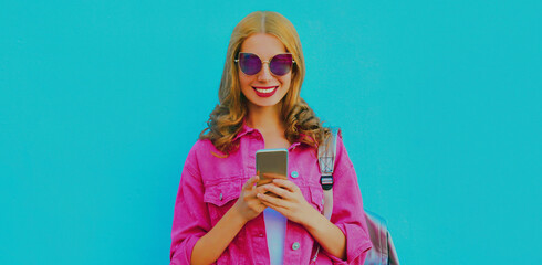 Portrait of stylish young woman with smartphone wearing a pink jacket, sunglasses over blue background
