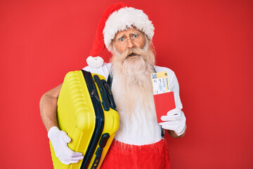 Old senior man wearing santa claus costume holding suicase and boarding pass in shock face, looking...