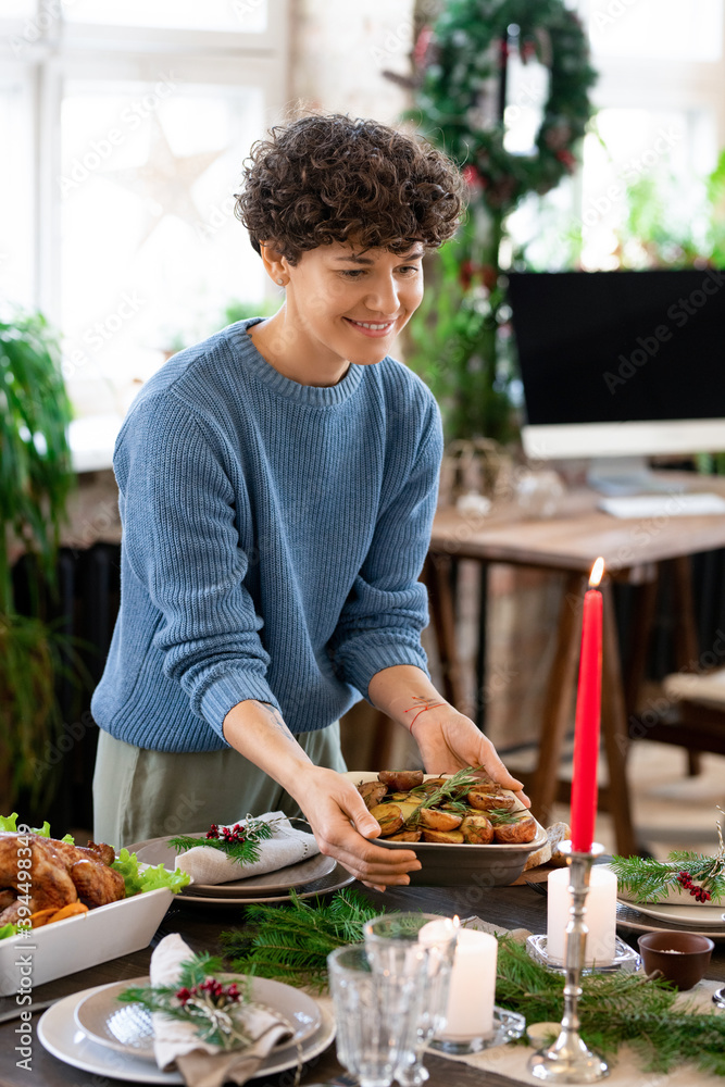 Wall mural Young smiling woman in blue knitted sweater and grey pants serving table