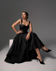 Elegant lady in black shiny ball gown and shoes sitting at white cibe with crossed legs and posing...