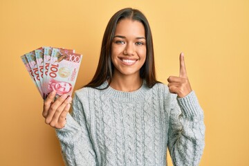 Beautiful hispanic woman holding 100 new zealand dollars banknote smiling with an idea or question pointing finger with happy face, number one