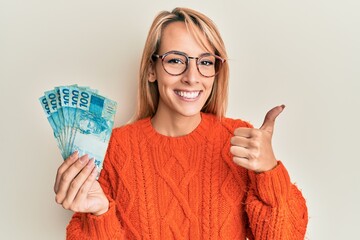 Beautiful blonde woman holding 100 brazilian real banknotes smiling happy and positive, thumb up...