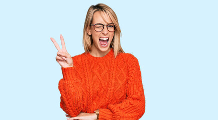 Beautiful blonde woman wearing casual clothes and glasses smiling with happy face winking at the...