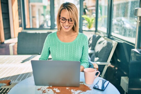 Young blonde woman smiling happy working using laptop sitting at the coffee shop terrace.