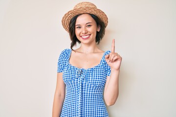 Young beautiful girl wearing summer hat smiling with an idea or question pointing finger up with happy face, number one