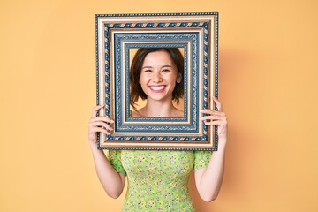 Young beautiful woman holding empty frame smiling with a happy and cool smile on face. showing...