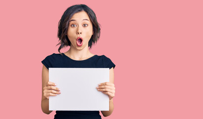 Young beautiful girl holding blank empty banner scared and amazed with open mouth for surprise,...