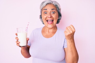 Senior hispanic woman holding glass of milk screaming proud, celebrating victory and success very excited with raised arms