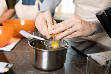 Hands of senior man in apron breaking egg into sieve over metallic pan by table