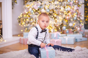A boy with Down syndrome and genetic pathology sits by the New Year tree and holds a box with a...