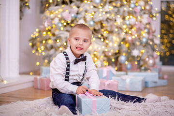 A boy with Down syndrome and genetic pathology sits by the New Year tree and holds a box with a...