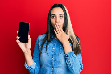 Young hispanic woman holding smartphone showing blank screen pointing thumb up to the side smiling happy with open mouth