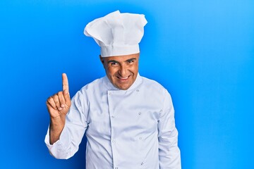 Mature middle east man wearing professional cook uniform and hat showing and pointing up with finger number one while smiling confident and happy.