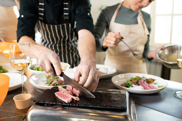 Hands of young male cooking coach chopping piece of smoked beef by table