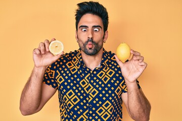 Handsome hispanic man with beard holding lemon making fish face with mouth and squinting eyes,...
