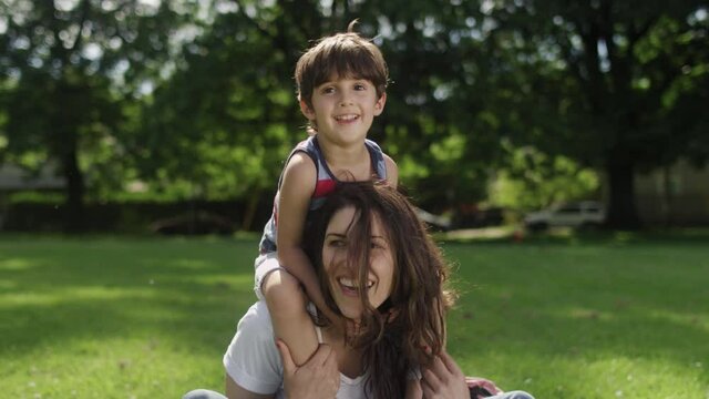A young boy crawls all over his mother's shoulders and head while sitting on the grass