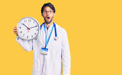 Handsome hispanic man wearing doctor uniform holding clock scared and amazed with open mouth for surprise, disbelief face