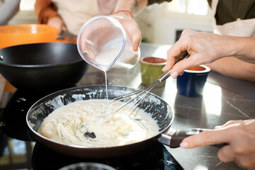 Hands of young man pouring milk into frying pan with flour while cooking