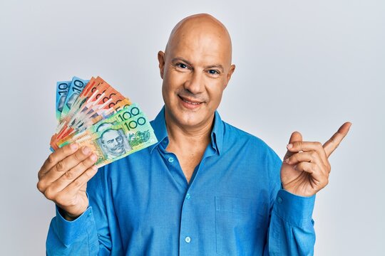 Middle age bald man holding australian dollars smiling happy pointing with hand and finger to the side