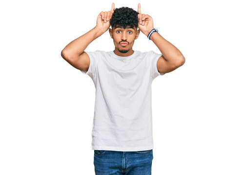 Young arab man wearing casual white t shirt doing funny gesture with finger over head as bull horns